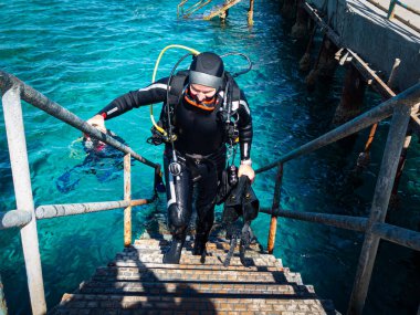 50 yaş üstü bir tüplü dalgıç sırtında ağır ekipmanlarla bir tüplü dalıştan dönen iskeleye doğru merdivenleri tırmanır.