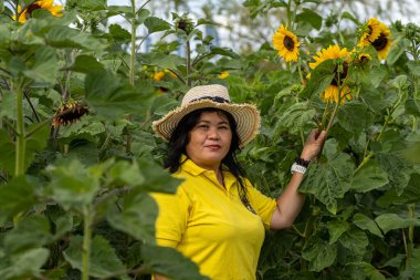 Orta yaşlı Asyalı bir kadının resmi, 50. yaş gününde, hasır şapkalı. Mavi gökyüzü arkaplanı