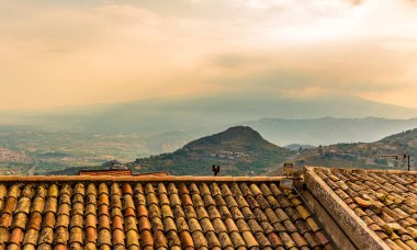 Castelmola, Sicilya 'nın çatıları boyunca batan güneşe ve yazın akşam sisi altındaki Etna Dağı' na doğru bir manzara.