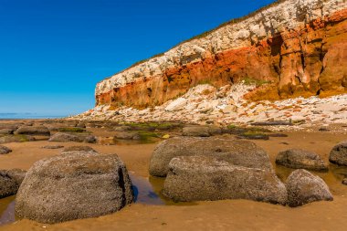 Midye kabuklu kayalar, değişik renklerdeki kayalar ve Old Hunstanton, Norfolk, İngiltere 'deki beyaz, kırmızı ve turuncu katmanlı kayalıkların tabanında kısmen yansıyan kaya havuzları.