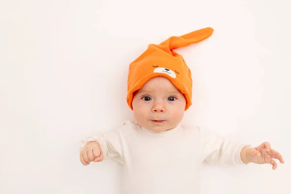 smiling baby girl in a white bodysuit in a bright orange hat on a white isolated background lies, portrait of a child 3-6 months old, space for text. A healthy happy child.