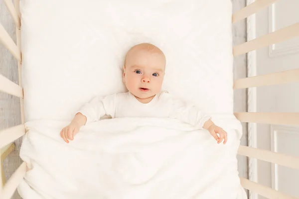 Portrait of a baby in a white crib. Interior of the children\'s room and bedding for children. A cute little girl is lying in her crib. The baby is Napping in the white bedroom. A healthy child in body-pajamas.