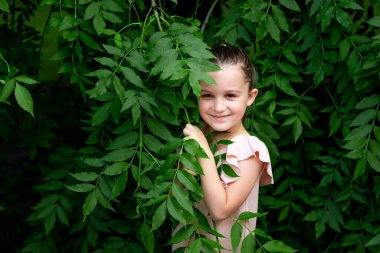 Gülümseyen kız 5-6 yaşlarında bir ağacın yanında duruyor, mutlu bir çocukluk, çocuk günü.