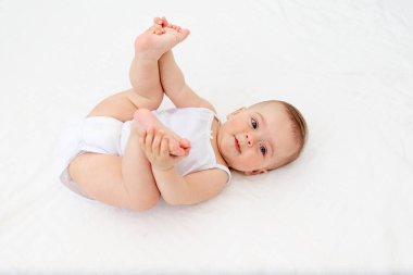 Bebek 6 aylık bebek beşikte sırt üstü uzanıyor ve bacaklarını tutuyor, kameraya bakıyor, bebek ürünleri konsepti.