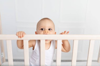 8 aylık bir bebeğin portresi. Çocuk odasında beyaz giysiler içinde duruyor ve yatağı kemiriyor. Bebek eşyaları konsepti.