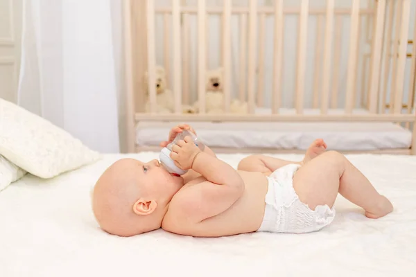 男婴8个月大 穿着尿布躺在白色的床上 脚上放着一瓶牛奶 婴儿食品的概念 婴儿从瓶子里喝水 发短信的地方 — 图库照片
