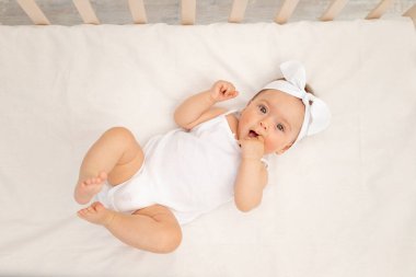 Bebek 6 aylık, ağzında parmaklarıyla bir çocuk odasındaki beşikte sırtına beyaz elbiseler giyip kameraya, bebek ürünleri konseptine bakarak yatıyor..