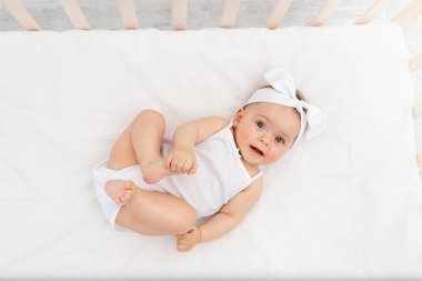 Bebek 6 aylık bebek beşikte yatıyor, sırtında beyaz elbiseler ve gülüyor, kameraya bakıyor, bebek ürünleri konsepti.