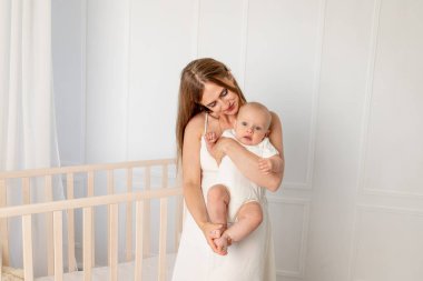 Genç ve güzel bir anne kızını altı ay bebek odasında tutuyor. Beşiğin yanında duruyor, anneler günü, mesaj için yer var.