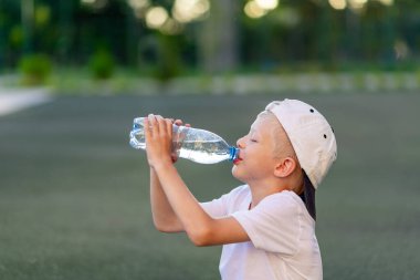 Spor üniformalı sarışın bir çocuğun portresi futbol sahasındaki yeşil çimlerde oturuyor ve bir şişeden, spor bölümünden su içiyor. Çocukların eğitimi, boş vakitleri..