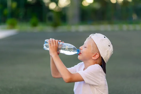 スポーツユニフォームを着た金髪の少年がサッカー場の緑の芝生の上に座りボトルやスポーツコーナーから水を飲んでいる姿が描かれています 子供のトレーニング 子供のレジャー — ストック写真