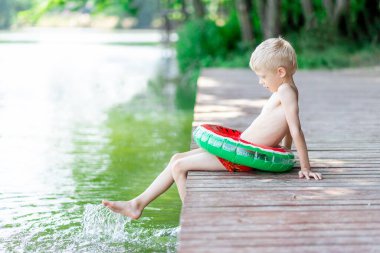Gülümseyen sarışın bir çocuk gölün kıyısındaki bir rıhtımda oturur ve sudan su sıçratır, bir çocuk yüzmeyi öğrenir, bir çocuk köyde dinlenir.