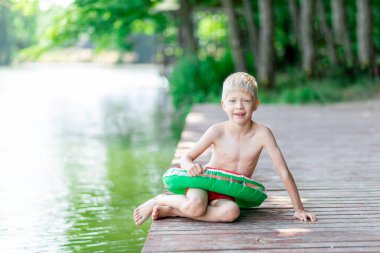 Gülümseyen sarışın çocuk göl kıyısındaki bir rıhtımda oturuyor, bir daire çiziyor, yüzüyor, bir çocuk yüzmeyi öğreniyor, köyde bir çocuk dinleniyor.