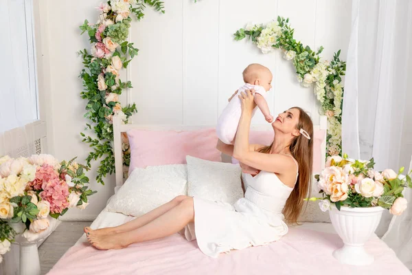 young beautiful mother holds her daughter a girl of 6 months in her arms lifting her up on a white bed, mother's day, place for text