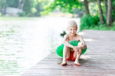 Gülümseyen sarışın çocuk göl kıyısındaki bir rıhtımda oturuyor, bir daire çiziyor, yüzüyor, bir çocuk yüzmeyi öğreniyor, köyde bir çocuk dinleniyor.