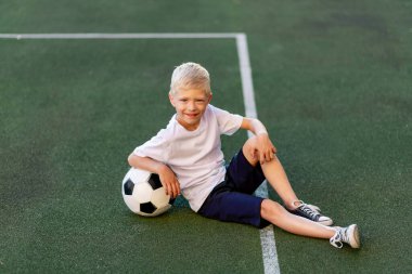 Spor üniformalı sarışın bir çocuk futbol sahasında futbol topuyla, spor bölümünde oturuyor. Çocukların eğitimi, boş vakitleri..