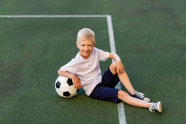 a blond boy in a sports uniform sits on a football field with a soccer ball, sports section. Training of children, children\'s leisure.