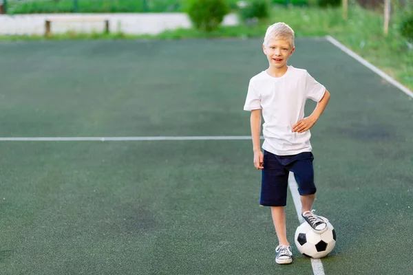a blond boy in a sports uniform stands on a football field with a soccer ball, sports section. Training of children, children's leisure.