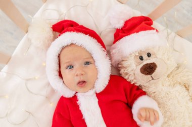 Noel Baba kostümü giymiş bir bebeğin Noel Baba şapkalı bir beşikte yattığı bir Noel fotoğrafı..