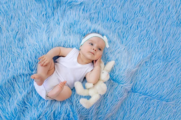 一个身穿白色紧身衣 面带微笑的孩子躺在蓝色毛皮的背景上 手里拿着一个玩具 望着别处 — 图库照片