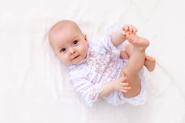 6 aylık küçük bir kız bebek beyaz bir elbise içinde hafif bir yatakta uzanıyor ve kameraya gülümsüyor. Bebek sırt üstü uzanıyor ve bacaklarını tutuyor.