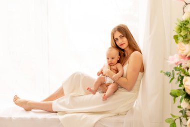 Genç bir anne, pencerenin önünde otururken bir çocuğu nazikçe kucaklıyor. Annesinin çocuğuna olan sevgisi ve bakımı.