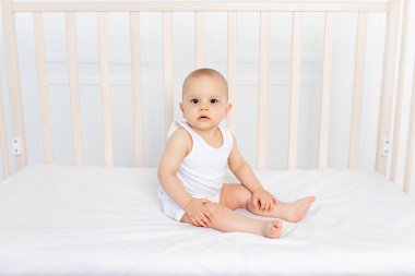 8 aylık bir bebek beşikte beyaz elbiseli bir çocuk odasında oturuyor ve bebeğin sabahında kameraya bakıyor.