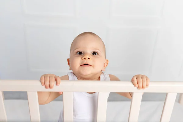 一个笑着的男婴的画像8个月大的男婴穿着白衣站在儿童房的婴儿床里 看着摄像机 早上的婴儿 婴儿产品的概念 — 图库照片