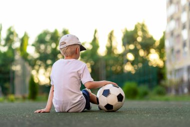 Spor elbiseli bir çocuk futbol sahasında yeşil çimlerde oturuyor. Sırtında futbol topu, gün batımında, dikiz manzarası, spor sayfası, antrenman..