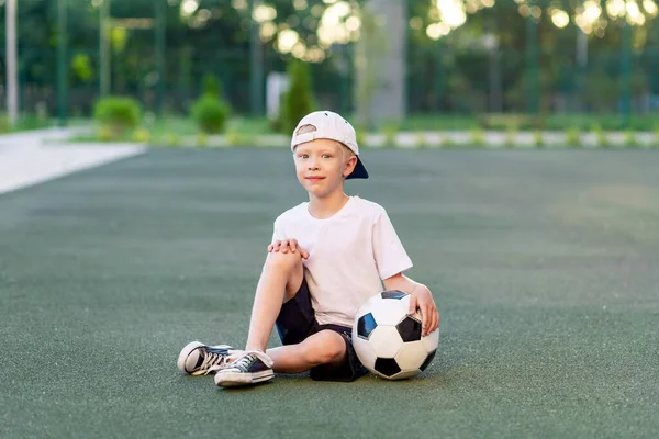a blond boy in a cap in a sports uniform sits on a football field with a soccer ball, sports section. Training of children, children\'s leisure.