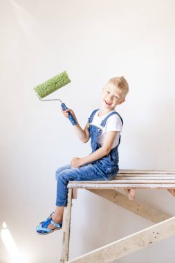 Gülümseyen bir çocuk inşaatçı, beyaz duvarları olan bir apartmanın merdiveninde oturur ve duvarları boyamak için bir yer tutar, yazı için bir yer, tamir konsepti.