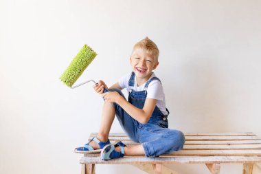 Gülümseyen bir çocuk inşaatçı, beyaz duvarları olan bir apartmanın merdiveninde oturur ve duvarları boyamak için bir yer tutar, yazı için bir yer, tamir konsepti.