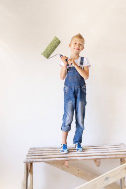 Sarışın bir çocuk, beyaz duvarlı bir apartman merdiveninin üzerinde duruyor ve elinde bir silindir, yazı için bir yer, tamir konsepti.