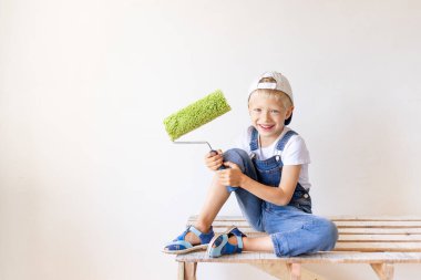 Gülümseyen bir çocuk inşaatçı, beyaz duvarları olan bir apartmanın merdiveninde oturur ve duvarları boyamak için bir yer tutar, yazı için bir yer, tamir konsepti.
