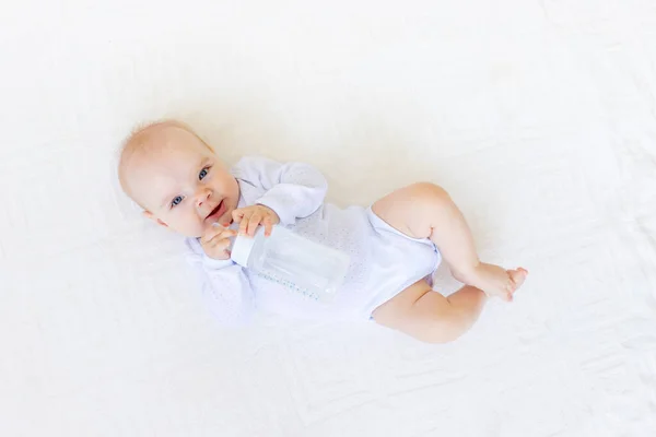 6个月大的小女孩 身穿白色的紧身衣 躺在家里的一张白色床上 抱着一瓶水和婴儿食品的概念 — 图库照片