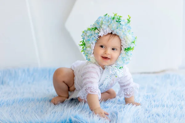 美しい女の子が花でできた帽子をかぶって笑顔で明るい部屋に青いふわふわの敷物の上に座っている — ストック写真