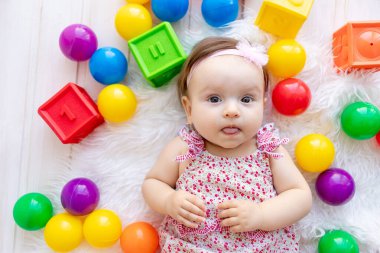 Güzel küçük bir kız bebek kırmızı giysiler içinde beyaz bir mindere uzanmış oyuncak topları ve küplerin arasında.