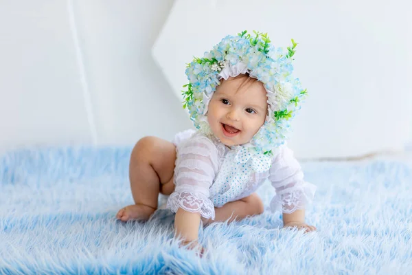 美しい女の子が花でできた帽子をかぶって笑顔で明るい部屋に青いふわふわの敷物の上に座っている — ストック写真