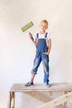 Sarışın bir çocuk, beyaz duvarlı bir apartman merdiveninin üzerinde duruyor ve elinde bir silindir, yazı için bir yer, tamir konsepti.