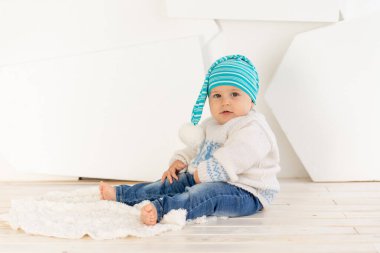 Küçük mutlu bir çocuk. Altı aylık, örgü örülmüş, kalın bir ceket ve parlak bir odadaki halının üzerinde bir şapka.