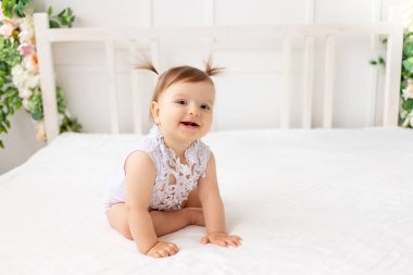 Altı aylık komik bir bebek, parlak güzel bir odada oturuyor, beyaz bir yatakta, dantelli bir elbise içinde kameraya bakıyor ve gülümsüyor.