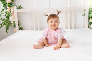 Neşeli kız altı aylık parlak güzel bir odada pembe elbiseli beyaz bir yatakta oturuyor ve gülümsüyor.