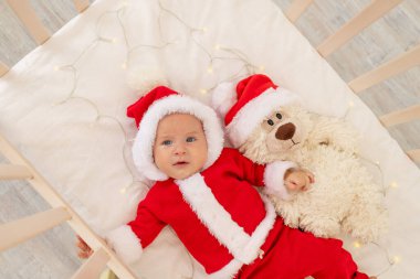 Noel Baba kostümü giymiş bir bebeğin Noel Baba şapkalı bir beşikte yattığı bir Noel fotoğrafı..