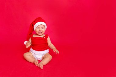 6 aylık Noel Baba şapkalı üzgün bebek kırmızı izole edilmiş fotofonda oturuyor ve ağlıyor, mesaj için yer, yeni yıl ve tatil konsepti.