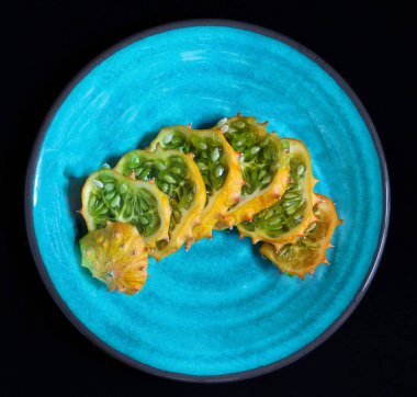 Parlak mavi bir tabakta dilimlenmiş kiwano meyvesi, ortalanmış ve güneş ışığıyla aydınlatılmış.