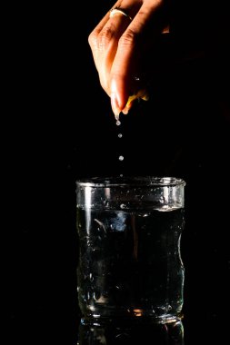 Karanlık bir arka planda yansıtıcı bir yüzeye yerleştirilmiş bir bardak su ve yukarıdan kesilmiş bir parça limon sıkılıyor ve suyu dökülüyor.