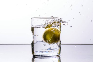 Beyaz arka planda yansıtıcı bir yüzeye yerleştirilmiş bir bardak su bardağa bir limon düştükten sonra sıçrıyor.