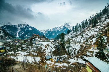 Çam ağaçları ve evleri olan bir dağda karla kaplı bir vadi manzarası. Bulutlu mavi gökyüzü