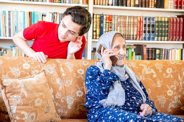 Happy arabic muslim grand mother and her son sitting together on sofa and using mobiles