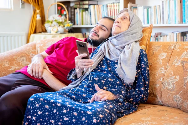 Happy arabic muslim grand mother and her son sitting together on sofa and using mobiles